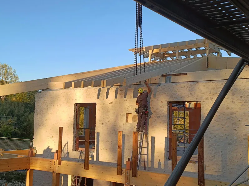 Charpentier fixant un chevron de la charpente bois au-dessus d'un mur en pierre, au soleil couchant